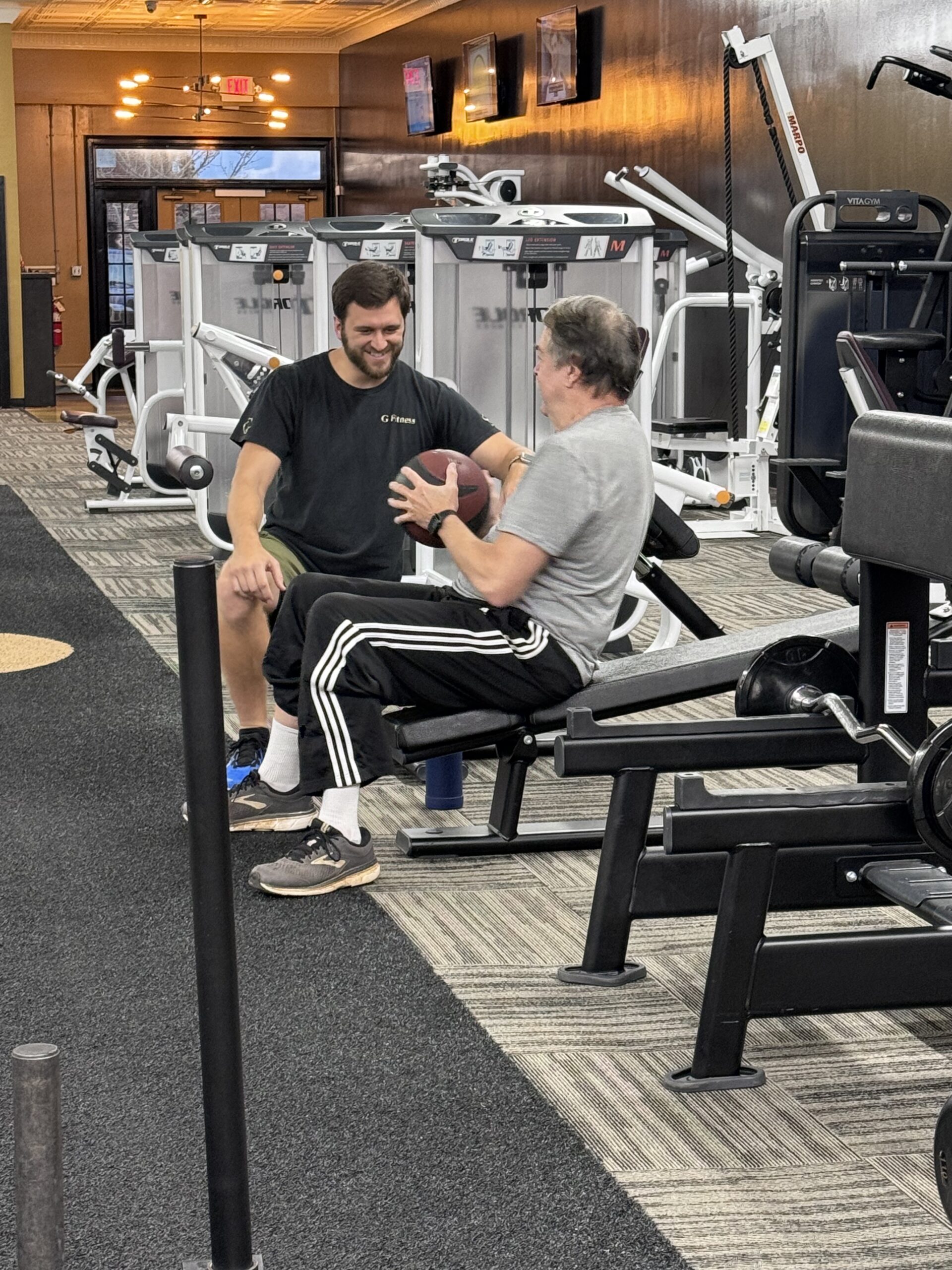 A trainer coaches a seated member holding a medicine ball at G Fitness