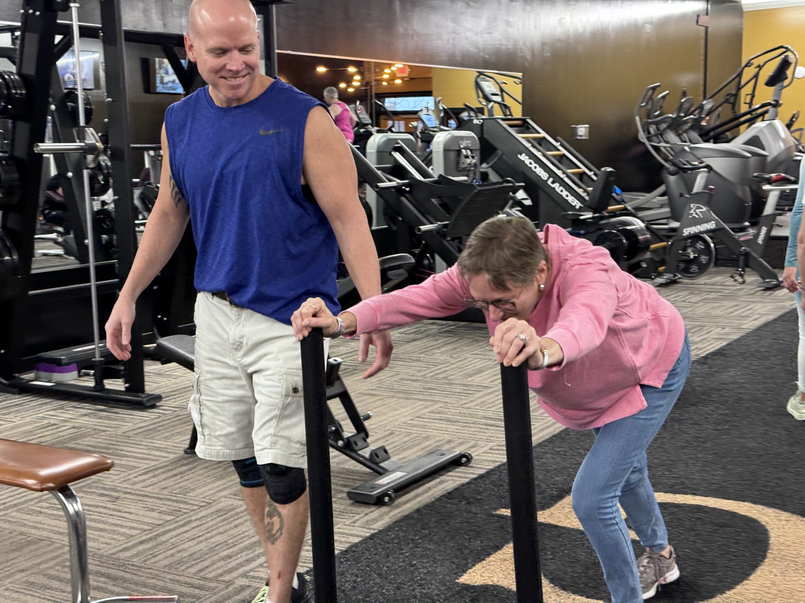 A member pushes a sled with a coach nearby at G Fitness