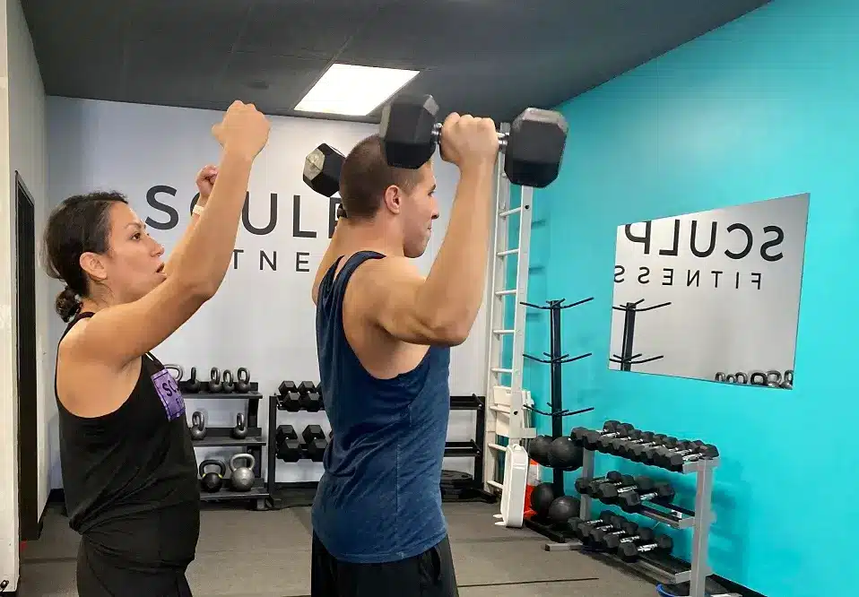 A Sculpt Fitness coach guiding a client through a dumbbell shoulder press